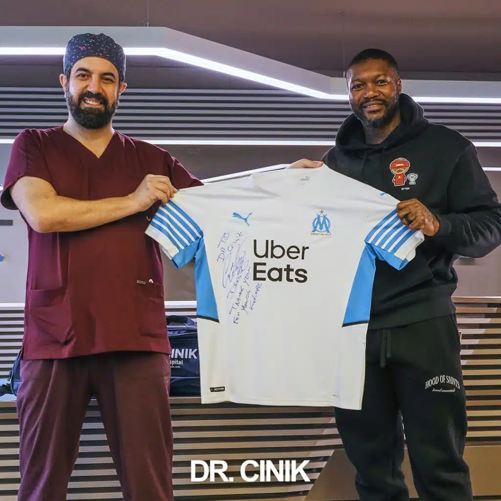 Dr. Cinik posing with former professional footballer Djibril Cissé holding a signed Olympique de Marseille jersey at Dr. Cinik's hair transplant clinic in Istanbul, Turkey